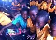 Three Ghanaian children, in orange school uniforms, in a classroom crowding around a computer.