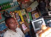 The young man, Martin Kakuru, in his store with a computer screen that he has repaired, and a pile of bread rolls he has made. 